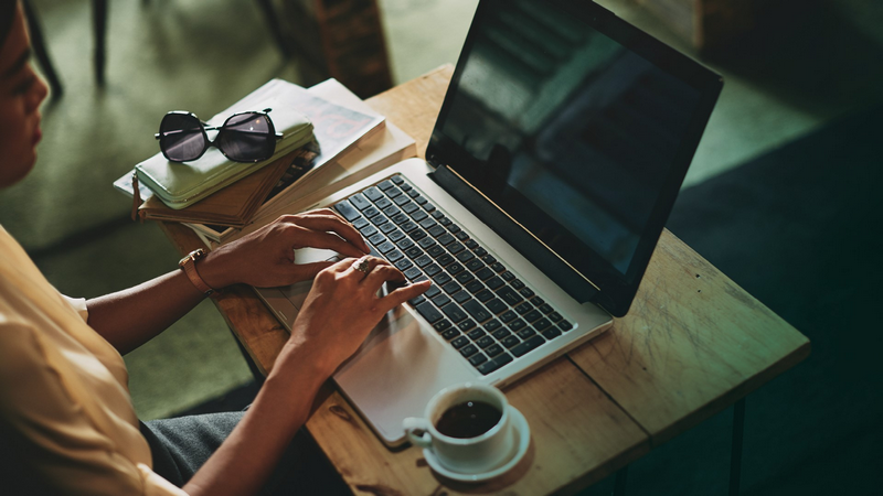 A person typing on a laptop with a coffee cup, books, and sunglasses nearby on a table.