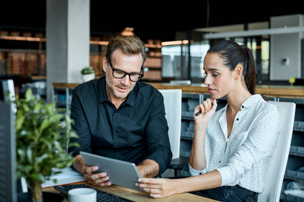 Two business professionals discuss strategies while looking at a tablet in a modern office setting.