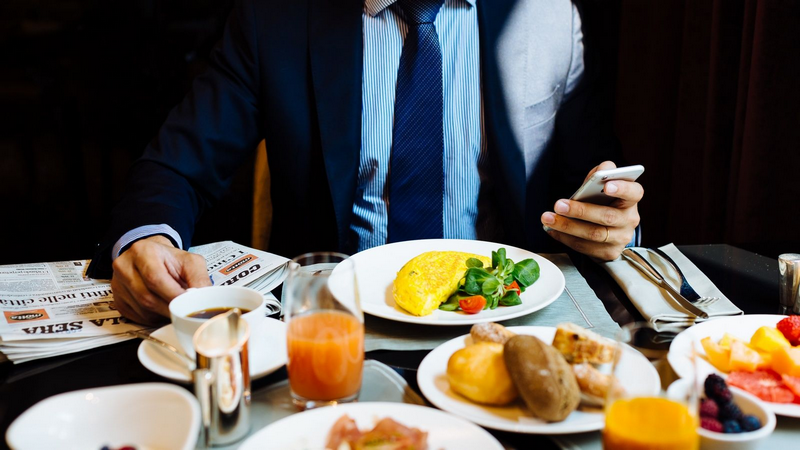 A person in a suit having breakfast while checking their phone and reading a newspaper.