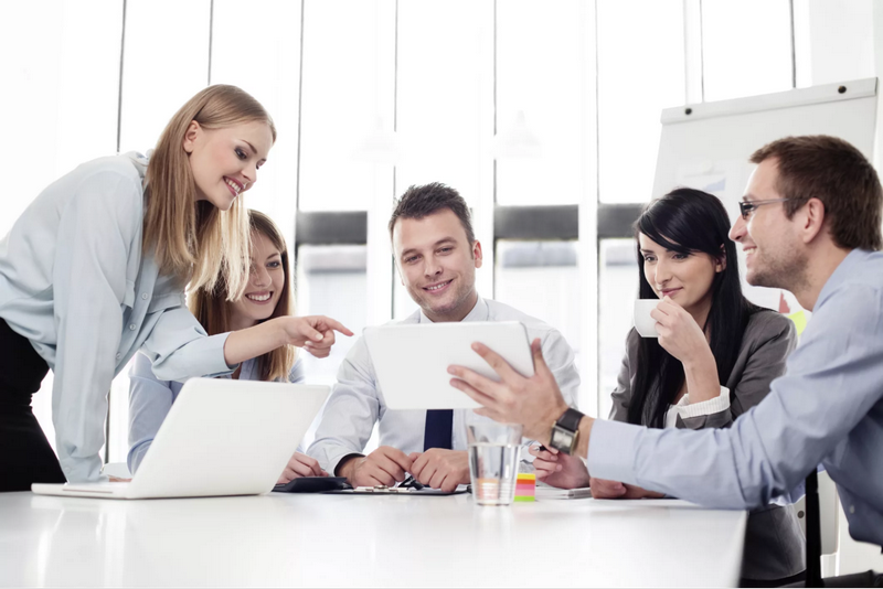 A group of five people in business attire are having a meeting around a table, looking at a tablet and smiling.