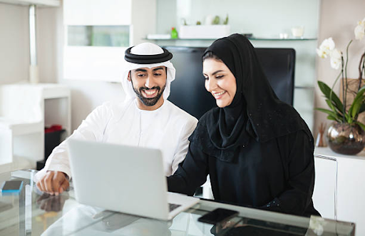 Two entrepreneurs, dressed in traditional attire, happily working together on a laptop in a modern office.